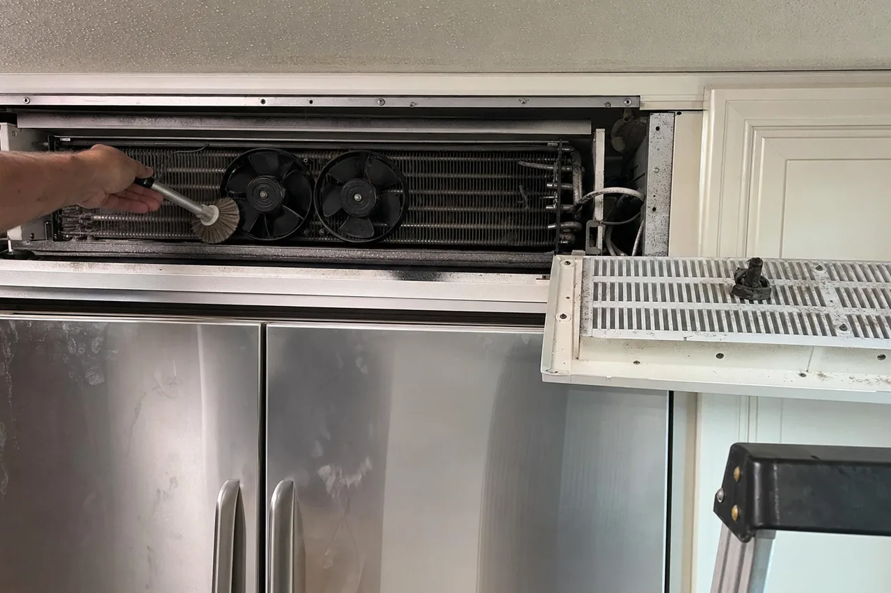 A brush being run through the condenser coil of a refrigerator with its vented grille panel swung down and both fans exposed