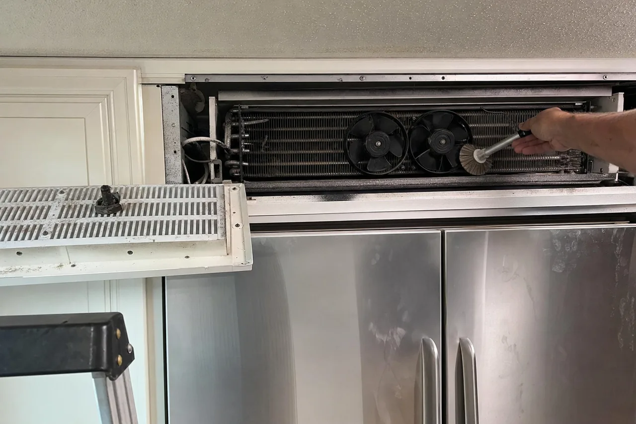 The condenser coil and twin fans of a refrigerator, grille panel hinged down and the coil part-cleaned