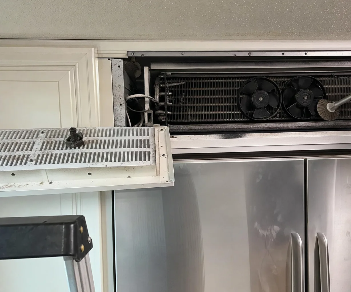 The condenser coil of a refrigerator part-way through a clean, brush in hand and the grille panel down