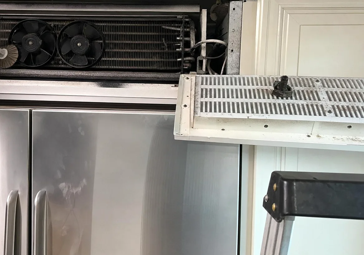 An opened refrigerator grille panel with the condenser coil and fans behind it