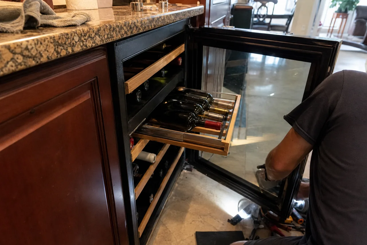 An undercounter wine cabinet under a speckled granite counter, glass door swung wide and a wood-fronted shelf of bottles pulled out while a technician kneels beside tools on the tile floor